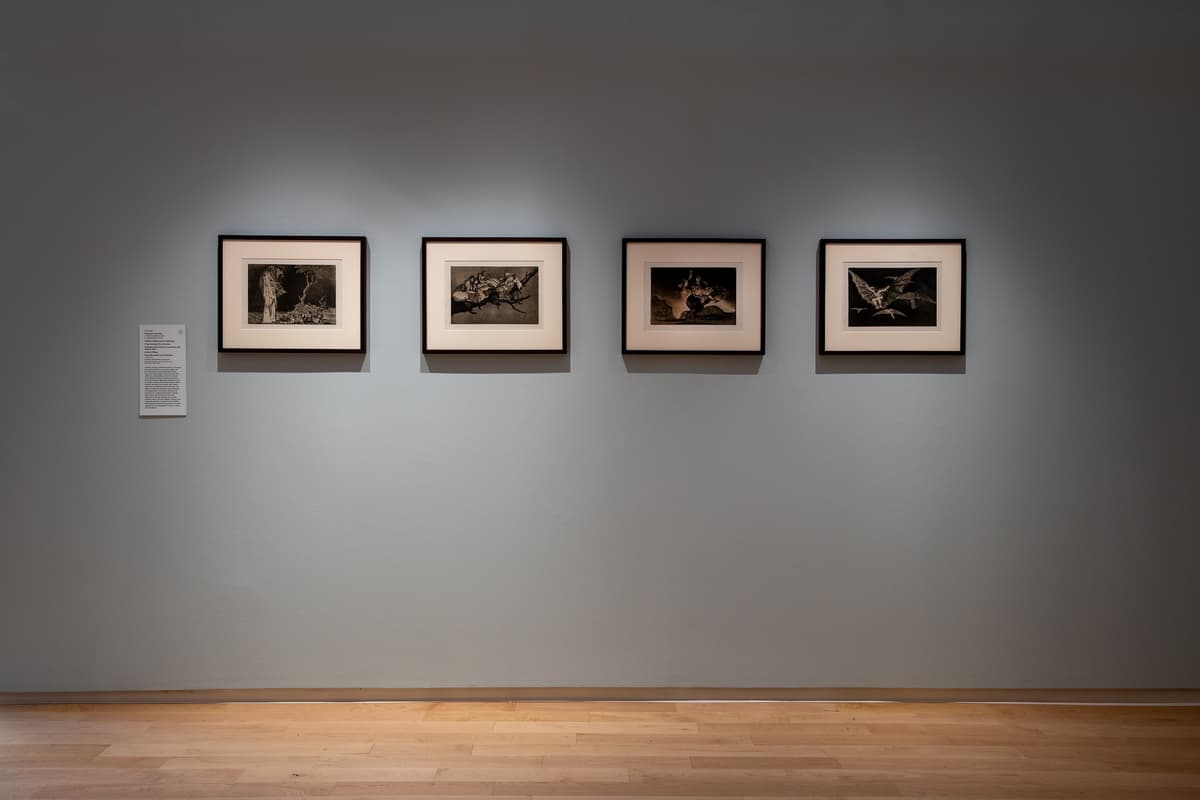 A symmetrical arrangement of four small, dark, framed etchings or prints hanging in a horizontal row on a neutral-toned gallery wall.