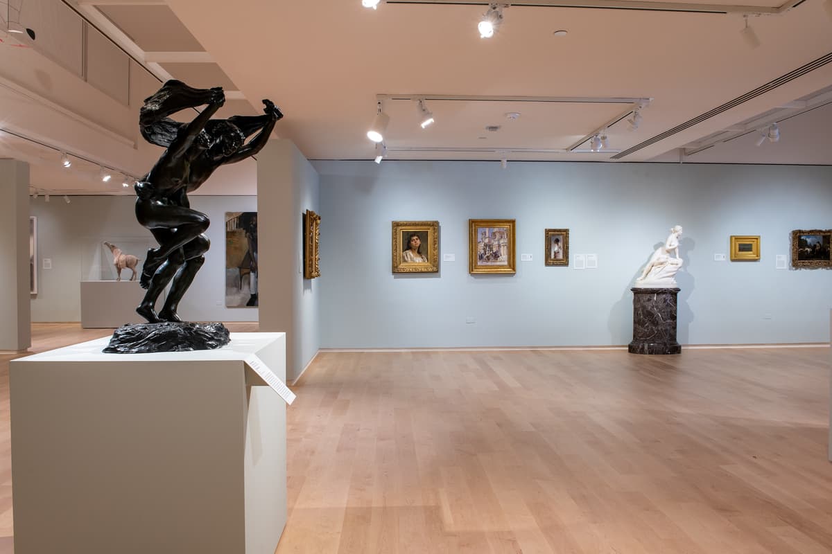 A wide-angle perspective of the museum interior, featuring a prominent black bronze sculpture of two figures in the foreground and various paintings and marble statues visible in the background.