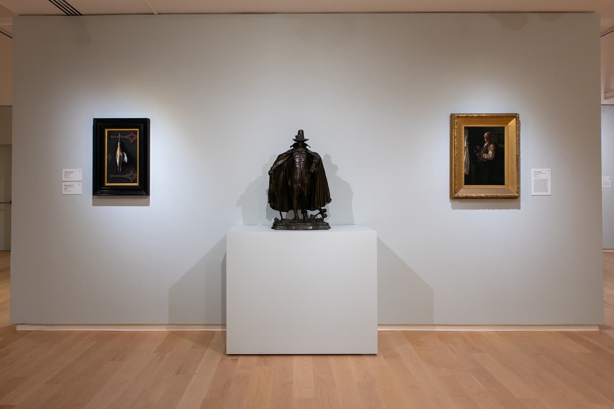 An eye-level shot of a gallery wall featuring a central bronze statue of a man in a wide-brimmed hat and cape, flanked by two framed paintings: a small still life on the left and a portrait on the right.