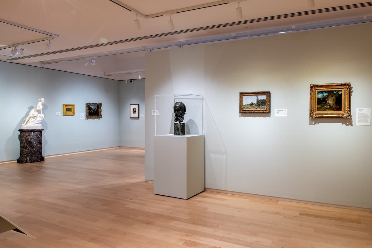 A view of a gallery corner featuring a bronze bust in a glass display case, several small framed landscape paintings, and a white marble statue in the distance.