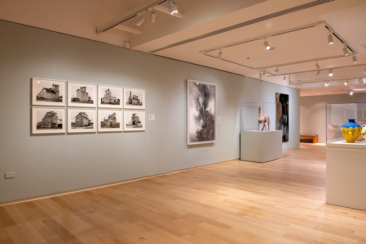 A wide view of a modern gallery space with light blue walls, featuring a grid of eight architectural photographs, a large vertical abstract print, and a sculpture of a horse in a glass case.