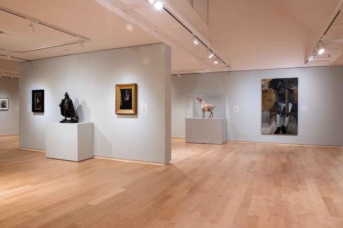 An installation view showing a dark bronze standing figure on a pedestal, a framed portrait, and a large ceramic horse sculpture in a glass case.