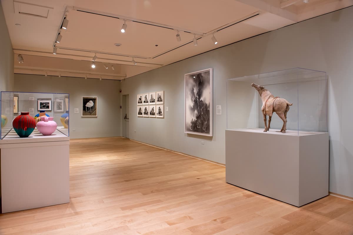 An installation view showing a dark bronze standing figure on a pedestal, a framed portrait, and a large ceramic horse sculpture in a glass case.