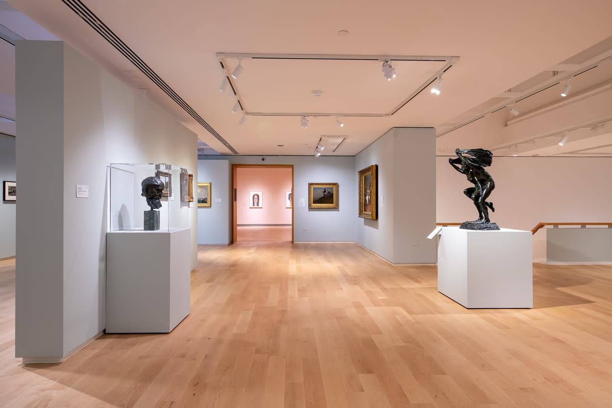 An eye-level view through the gallery toward a doorway, showing a marble bust in the foreground and a bronze sculpture of a figure carrying another.