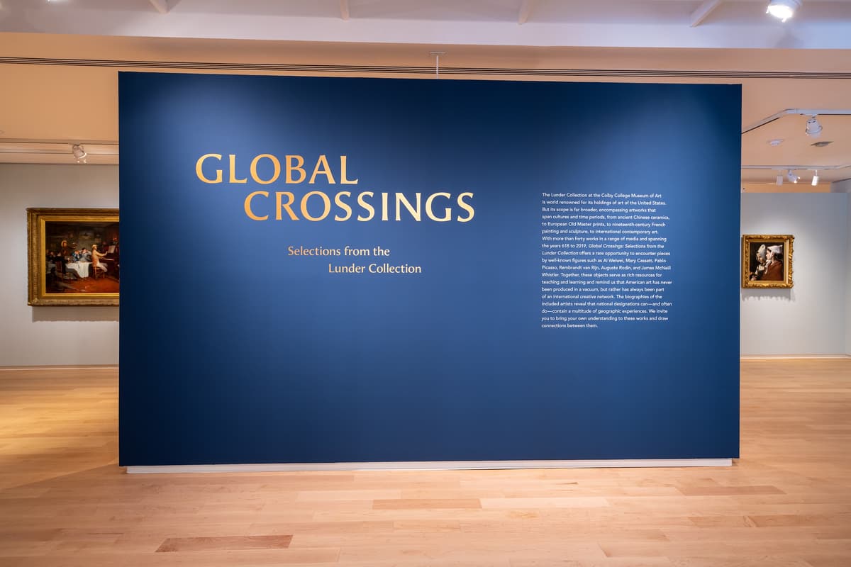 A large, dark blue exhibition title wall that reads "GLOBAL CROSSINGS: Selections from the Lunder Collection" in gold lettering.