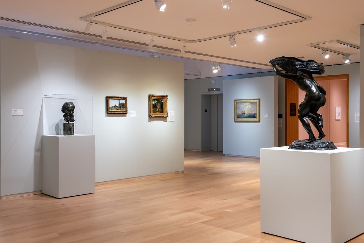 An angled view of an art gallery showing several framed landscape paintings on light-colored walls and two bronze sculptures: a bust in a glass case and a dynamic figure in motion on a white pedestal.