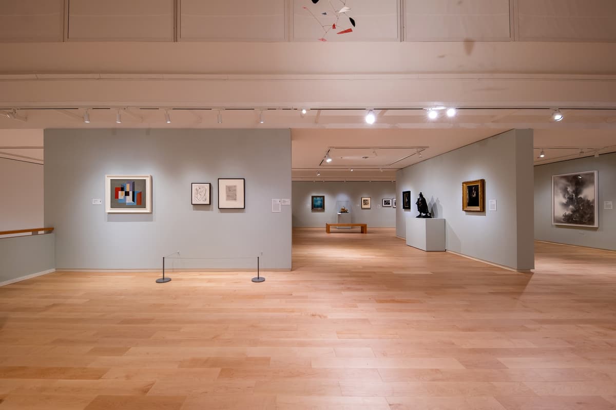 A high-angle view of the gallery space showing a clean layout of framed art, white pedestals, and a colorful mobile hanging from the ceiling.