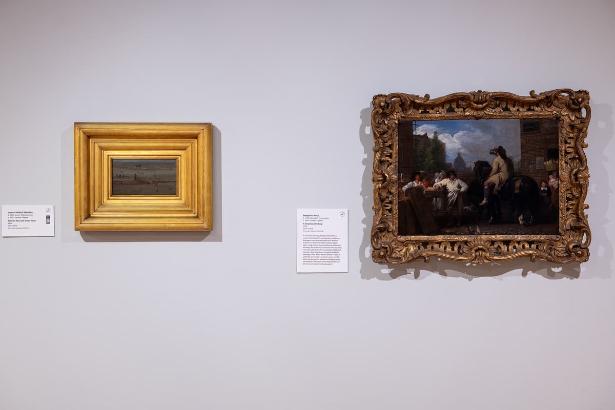 A close-up of two ornate gold-framed paintings on a light grey wall: a small, horizontal landscape and a larger scene of a street with a horse and rider.