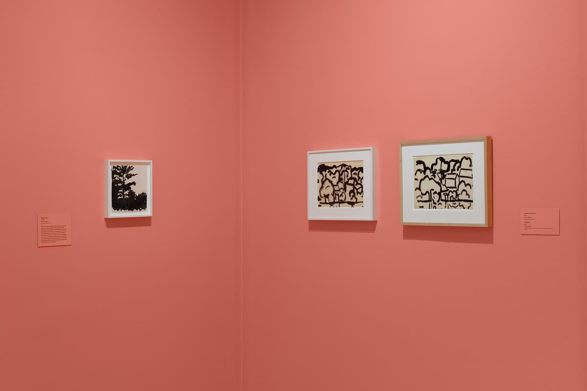 A view of a corner inside a gallery with coral-pink walls and light wood flooring. On the left wall hangs a single, small framed ink drawing of a dark tree silhouette. On the right wall hang two framed landscape drawings rendered in thick, bold black ink lines depicting trees and houses. A small pink description card is mounted next to each artwork grouping.