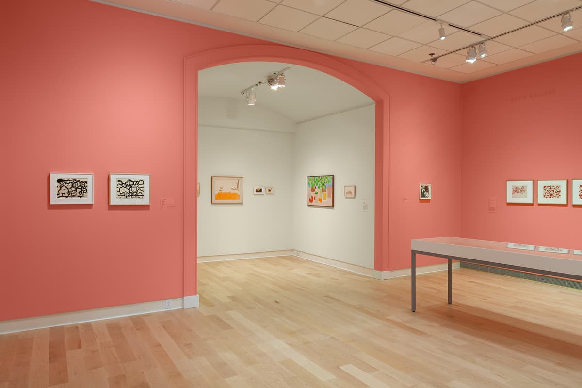 A wide view of a gallery space with coral-pink walls and a light hardwood floor. A large open archway in the center leads into an adjacent white-walled gallery room displaying several colorful framed artworks. To the left of the archway, two framed black ink drawings hang on the coral wall. To the right, a long glass-topped display vitrine sits on the floor beneath overhead track lighting.