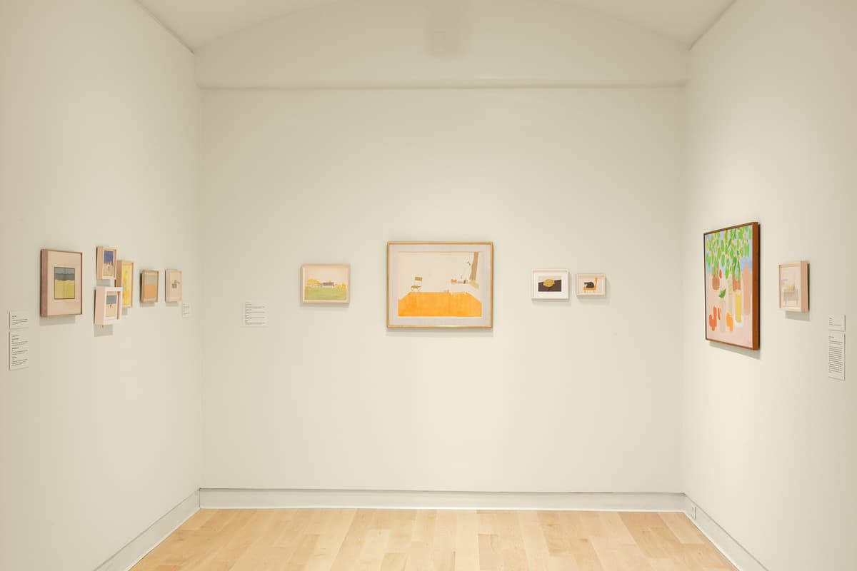 A straight-on shot of a small, white-walled gallery room with light hardwood floors. The back wall features four small framed artworks, including a central horizontal piece with a bright orange lower half depicting an interior space. The left wall displays a row of six small framed minimalist watercolor and paper collages. The right wall features a larger colorful painting of green foliage and a small framed interior sketch.