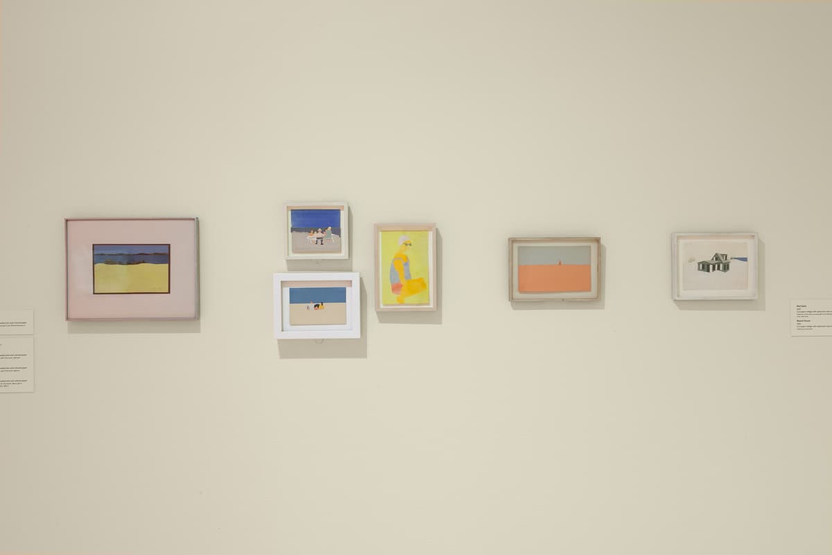 A close-up, horizontal arrangement of five small framed artworks on a white gallery wall. From left to right: a horizontal landscape with a yellow field and dark blue water; a vertical stack of two small beach scenes; a vertical yellow portrait of a person in a swimsuit; a minimalist horizontal landscape with a pink sky and orange ground; and a small horizontal collage of a green house against a white background. White description labels are visible on the far left and right.
