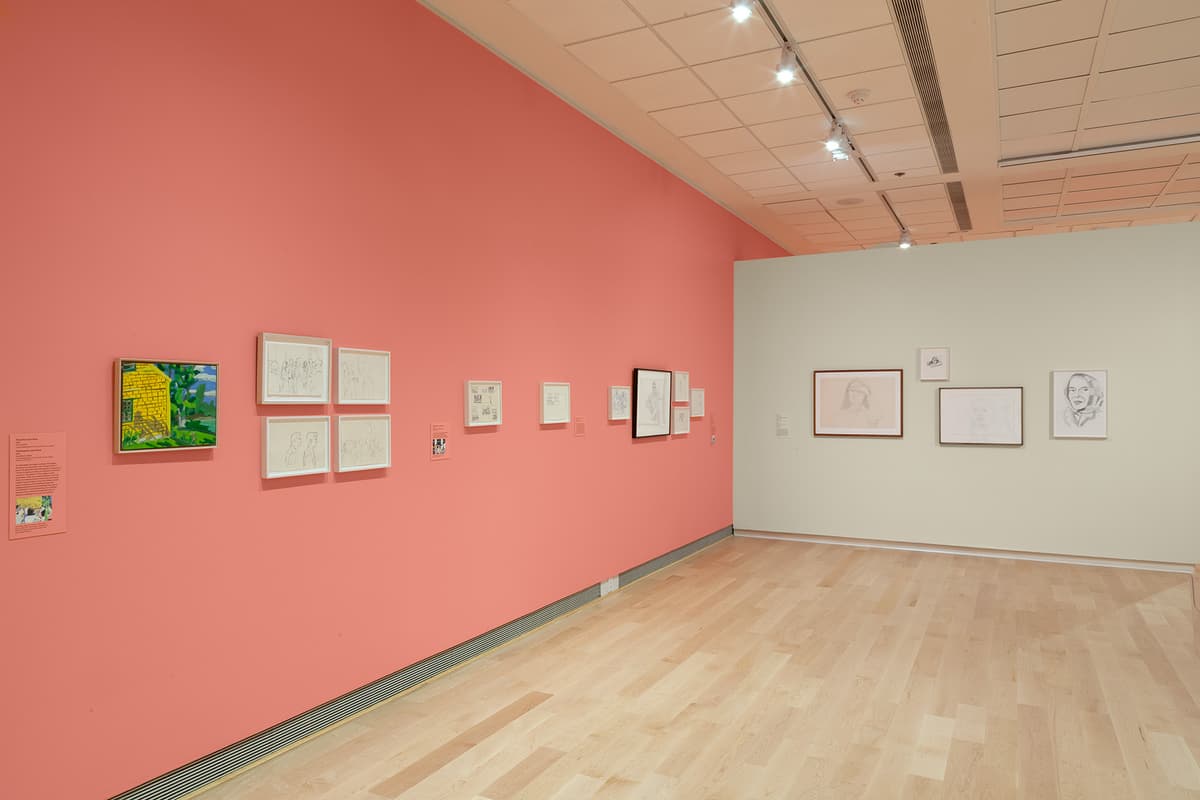 A view down a long coral-pink gallery wall that transitions to an off-white back wall. The coral wall on the left displays a small, colorful square painting of a yellow house next to green trees, followed by several small white-framed sketches arranged in pairs or small groups. The white wall in the background features three medium-sized framed portrait sketches.