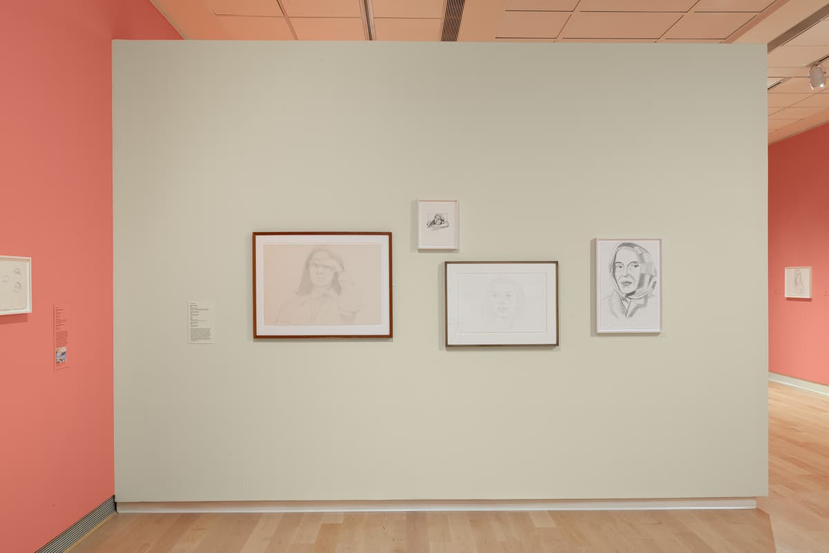 A straight-on view of an off-white freestanding gallery wall displaying four framed portrait sketches. On the left is a larger brown-framed sketch of a person looking forward with a hand to their forehead. To its right are three smaller sketches framed in white: one small landscape sketch on top, a horizontal portrait below it, and a vertical portrait of a woman wearing a headscarf on the far right. A coral-pink wall is visible on the left and far right background.