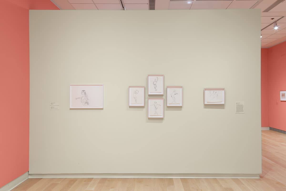 A straight-on view of the off-white freestanding wall showcasing five framed line drawings of dancers in motion. The artwork on the left is a larger horizontal frame showing a dancer with arms extended. The central group consists of four small, vertically oriented white frames arranged in a cross-like formation, each capturing a different balletic pose. A text block label is mounted on the far right.