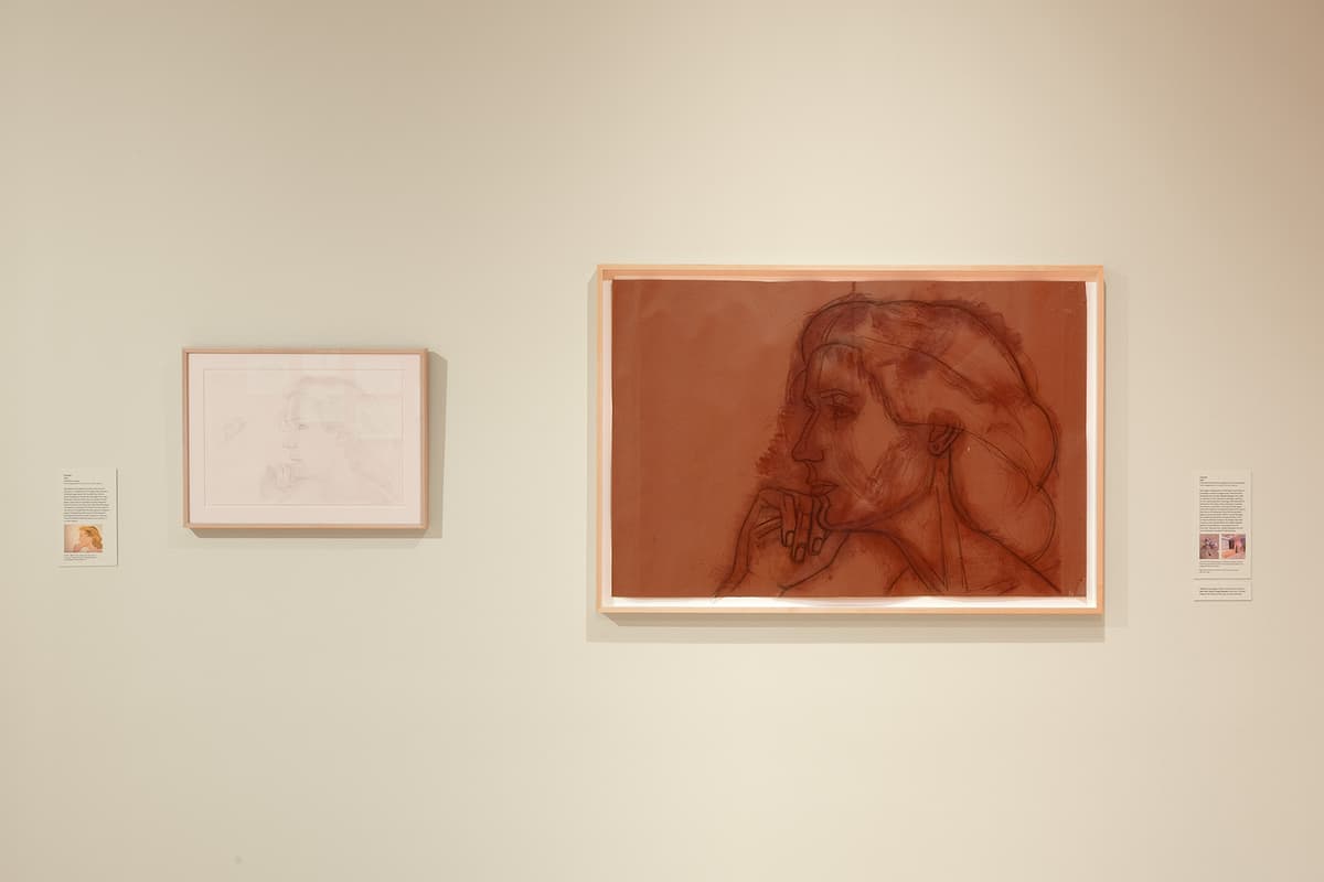 A close-up view of two artworks on an off-white wall. On the left is a small horizontal sketch of a woman in profile with a text label beside it. On the right is a large horizontal artwork featuring a bold, expressive charcoal or dark ink portrait sketch of a person resting their chin on their hand, set against a solid reddish-brown, terracotta-colored paper background.