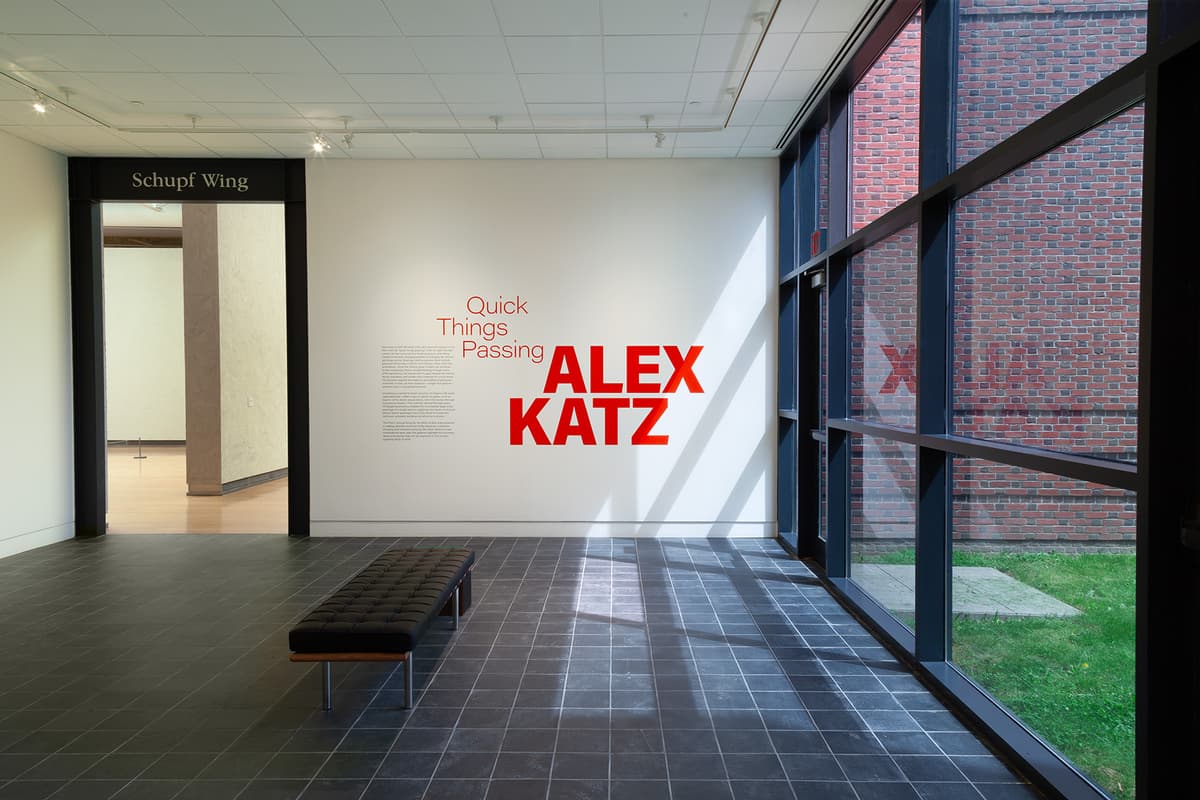 An exhibition entryway featuring a large white wall with vinyl text that reads "Quick Things Passing ALEX KATZ" in vibrant orange and red lettering, accompanied by an introductory wall text paragraph. To the right, a large grid window overlooks an outdoor brick wall and a patch of green grass. A long, black tufted leather bench sits on a dark grey tiled floor, and an open doorway on the left labeled "Schupf Wing" reveals an adjoining gallery room.
