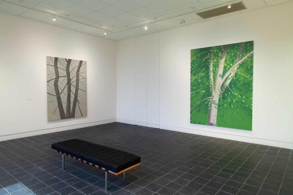 A wide angled view of a gallery corner with dark grey floor tiles and white walls. A black tufted leather bench rests on the floor in the foreground. The left wall features a large vertical painting of bare, dark tree branches against a muted gray sky. The right wall displays a large, vibrant vertical painting of a tree trunk surrounded by lush green summer foliage.