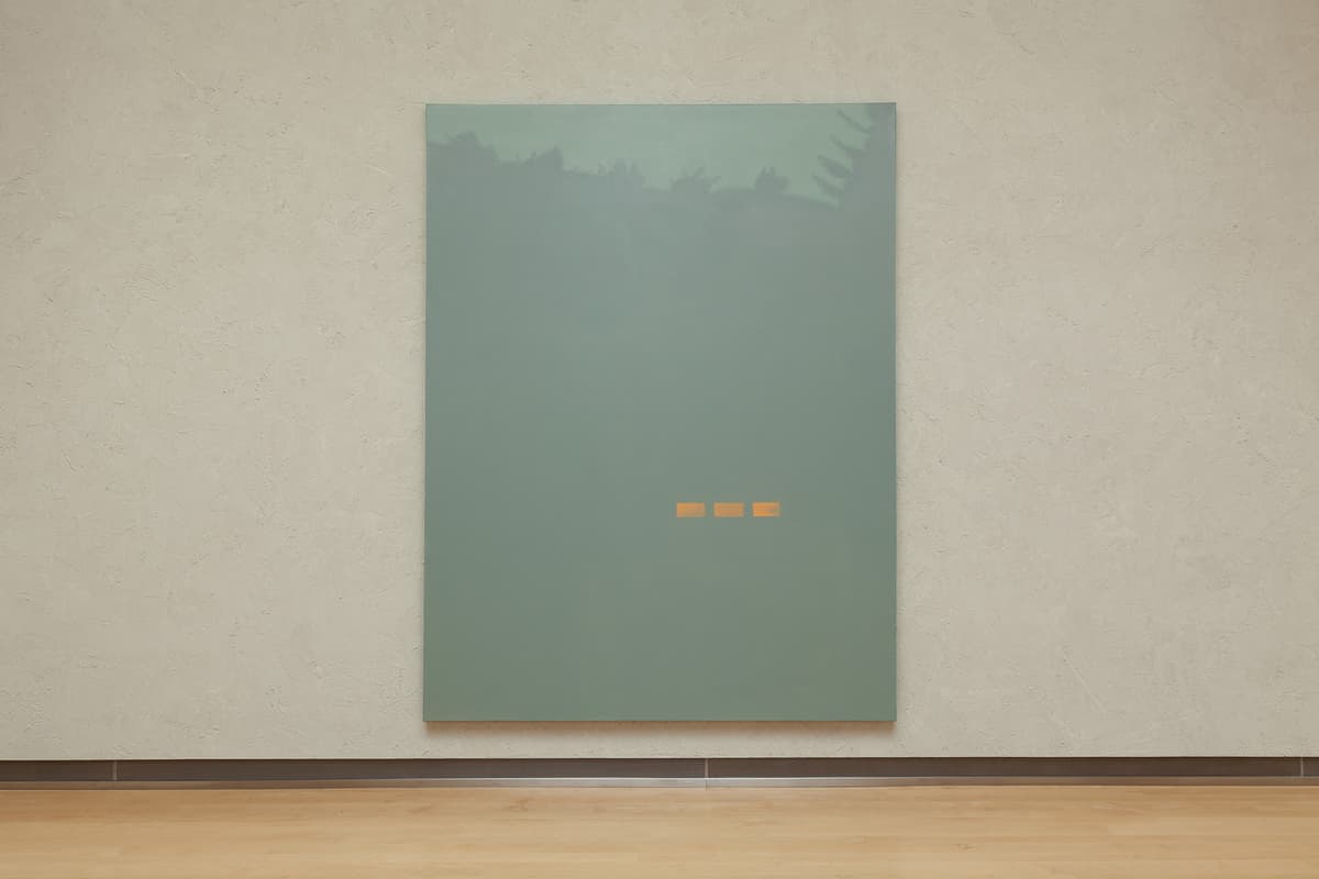 A straight-on view of a large vertical painting centered on a textured, light beige gallery wall above a light hardwood floor. The painting features a solid, minimalist field of muted sage green with three small, horizontal orange rectangular dashes arranged in a neat line near the lower center.