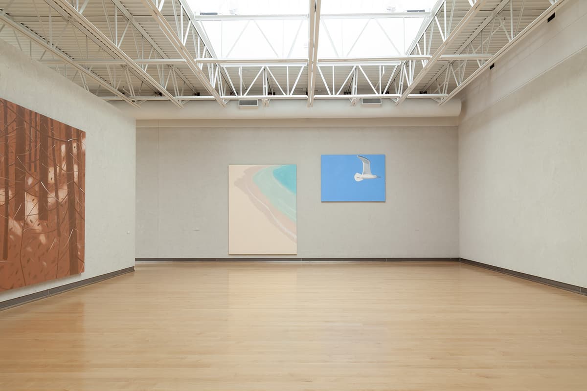 A spacious view of a well-lit gallery room under an open white metal truss ceiling with skylights. The light hardwood floor reflects the overhead light. The far wall displays two large paintings side by side: one depicts an abstract shoreline with soft turquoise water and beige sand, and the other shows a single white seagull soaring through a solid bright blue sky. A portion of a brown forest painting is visible on the far left.