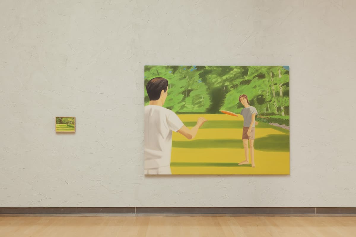 A straight-on view of a textured cream-colored wall showing two paintings. On the left is a very small, horizontally framed landscape painting. To its right is a large, horizontal painting depicting two figures playing frisbee on a vibrant yellow and green lawn, with a figure in a white shirt seen from behind in the foreground throwing a orange disc toward a figure standing in the distance.