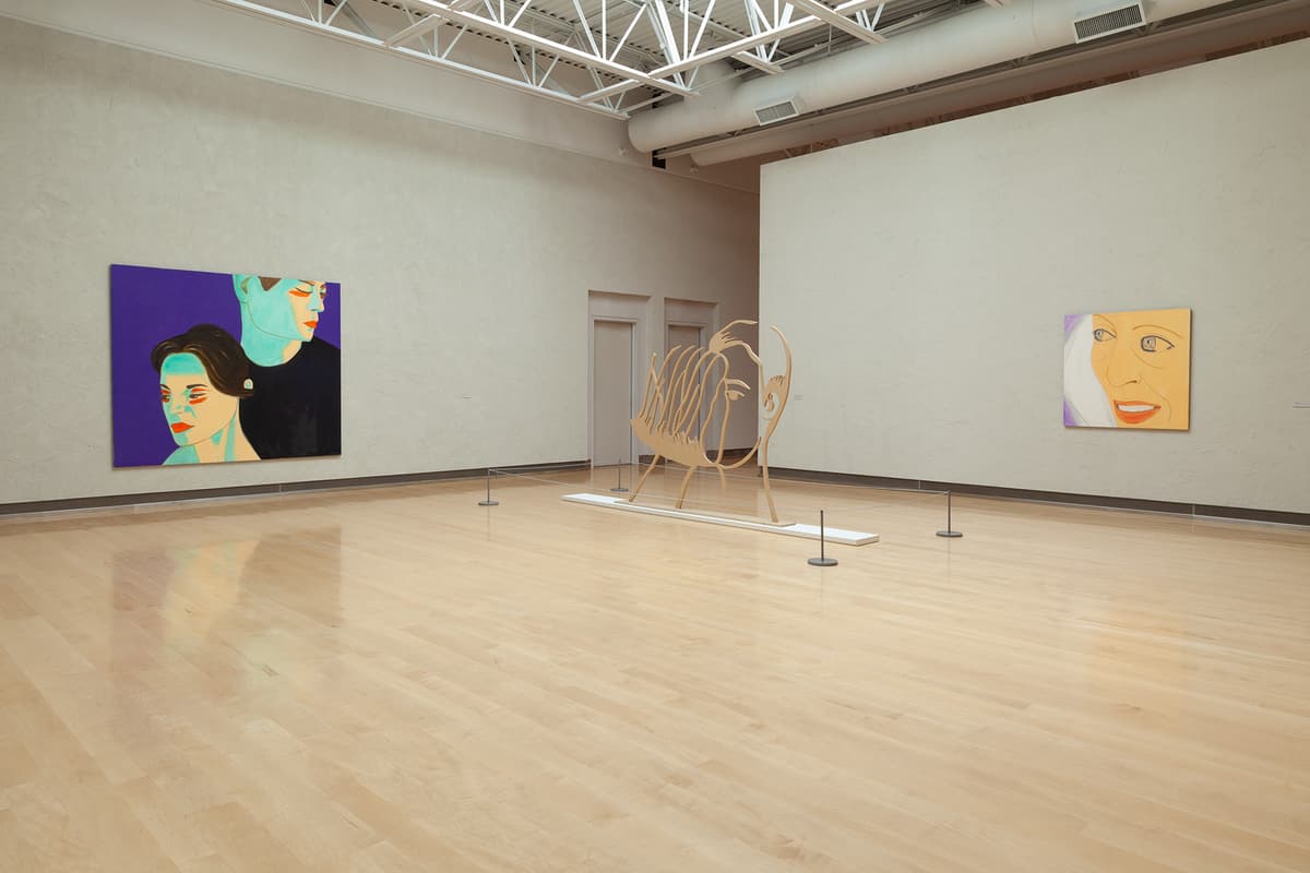 A wide-angle view of an expansive gallery room with a light hardwood floor. On the far left wall hangs a large horizontal portrait painting of a man and a woman in cool blue-green skin tones against a dark purple background. In the center of the room, a large freestanding wooden cutout sculpture of a stylized face with flowing hair is cordoned off by a low stanchion wire. To the right, a smaller square painting shows a close-up of a smiling woman's face on a yellow background.