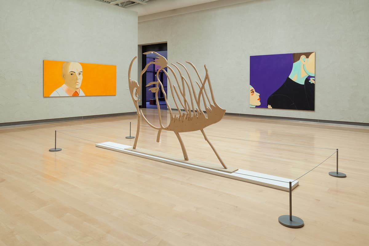 An alternate view of the gallery room featuring the freestanding wooden cutout sculpture of a face from behind, secured by low stanchions. In the background on the left wall, a horizontal painting features a close-up portrait of a bald man against a bright orange backdrop. On the right wall, a large vertical painting shows a close-up profile of a woman looking toward a man's shadowed figure against a deep purple background.