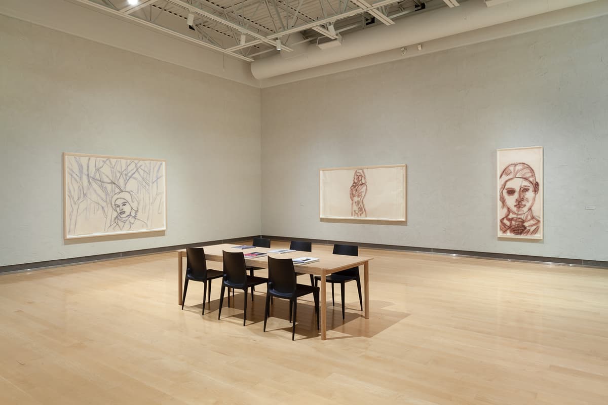 A wide shot of a gallery corner featuring a light wooden reading table surrounded by six black chairs in the center of a polished light hardwood floor. The textured walls display three large, loosely sketched charcoal portraits in simple light wood frames: one on the left showing a figure against abstract trees, one in the center depicting a standing woman, and a vertical one on the right showing a close-up face.