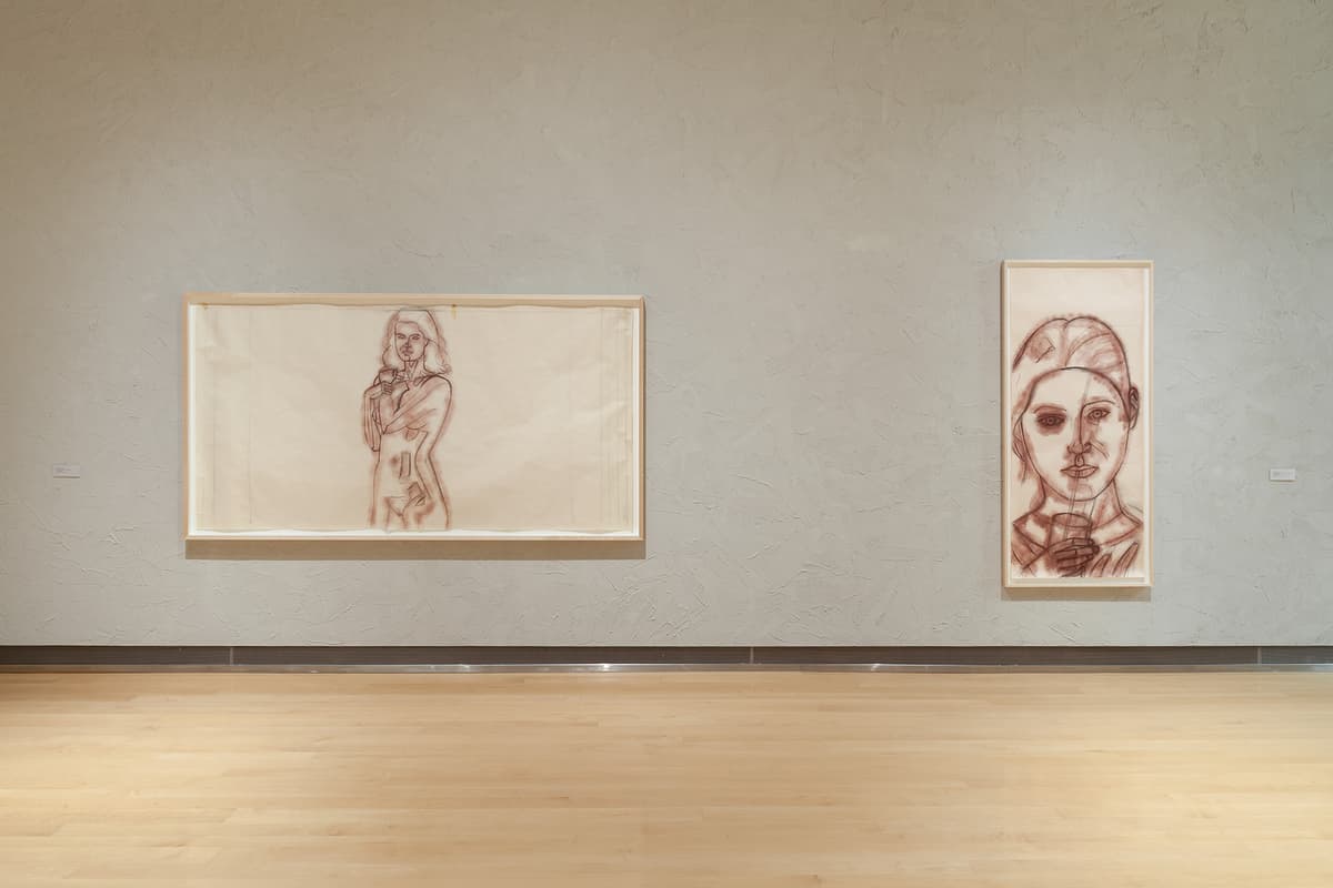 A straight-on view of a textured gray gallery wall displaying two large sketches in light wood frames above a hardwood floor. On the left, a wide horizontal sketch rendered in brown lines depicts a full-body figure of a woman holding a cup. On the right, a narrow vertical frame contains a large close-up portrait sketch of a person's face sipping from a straw.