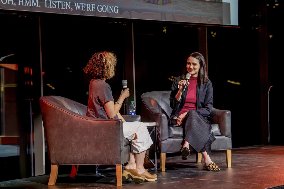 Two individuals seated on stage holding microphones talk to one another.