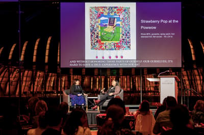 Two individuals seated on a stage with microphones. An audience is seated in front of them and behind them, a presentation is projected with a work of art titled "Strawberry Pop at the Powwow."