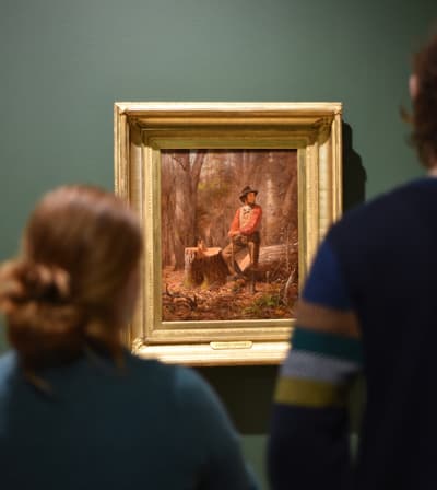 Two individuals look at a figurative painting in a gold frame hung on a dark green wall. It depicts an single man in a red coat and black hat seated in the woods.