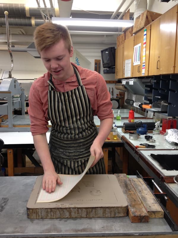 A medium shot shows a young man with short blonde hair and a light complexion, wearing a peach-colored button-down shirt and a black and white striped apron. He is standing in a printmaking studio, leaning over a large, flat, rectangular stone block on a metal table. He is carefully lifting a sheet of paper from the surface of the stone with his right hand while pressing down on the other side with his left hand to reveal a print. The print on the stone features two small sketches of heads and some faint text. The background is a cluttered workshop with wooden cabinets, various printmaking tools like rollers and ink tins, and industrial pipes along the ceiling. The lighting is bright and even, typical of a studio environment.