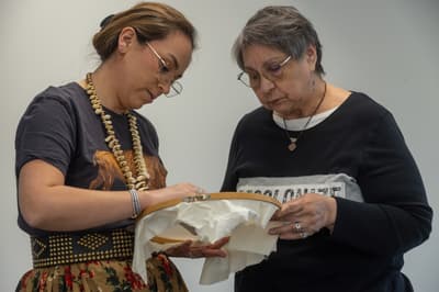 Two women wearing glasses lean in close to examine a piece of white fabric held in a wooden embroidery hoop.