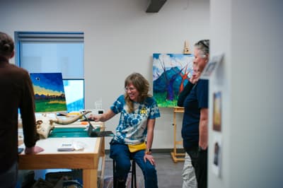 Individuals gathered in an artist studio, with vibrant landscape paintings in the background. One person is seated on a stool and using a tablet to show something to two people standing nearby.