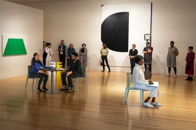 In a spacious art gallery, two performers sit at a small round table on the left, while a third performer in all-white clothing sits alone on the right holding a sketchbook. A small audience of museum-goers stands against the back wall, framed by large-scale geometric paintings in green, black, and white.