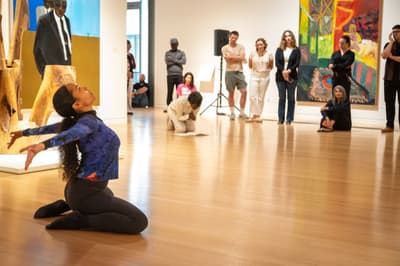 Performance artist on her knees in a gallery in front of an audience casts her gaze upward and spreads her arms out behind her