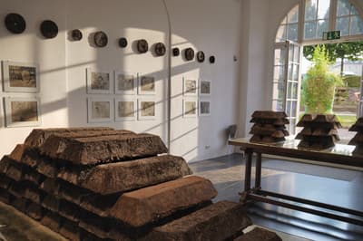 An interior gallery space featuring Claire Pentecost's "Soil-erg" installation. Large, dark, rectangular bricks made of soil are stacked in a pyramid shape in the foreground, while smaller stacks sit on a wooden table. The background wall displays circular soil discs and framed currency-like prints, illuminated by dramatic sunlight streaming through a large arched window.