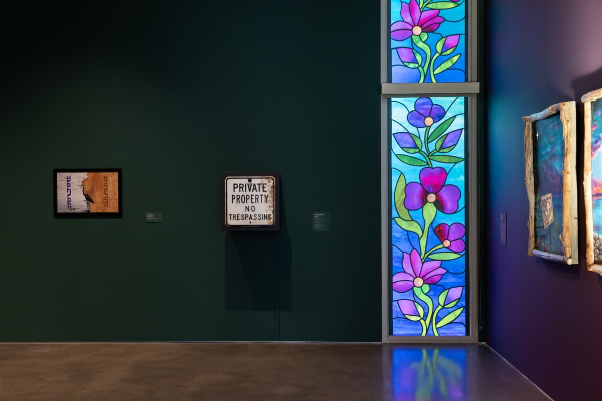 Gallery installation featuring a dark green wall. A "Private Property No Trespassing" sign and a small framed artwork are hung adjacent to a vibrant, vertical stained-glass window with a purple floral pattern. The adjacent wall is painted a purple-toned wall and two framed works are in partial view.