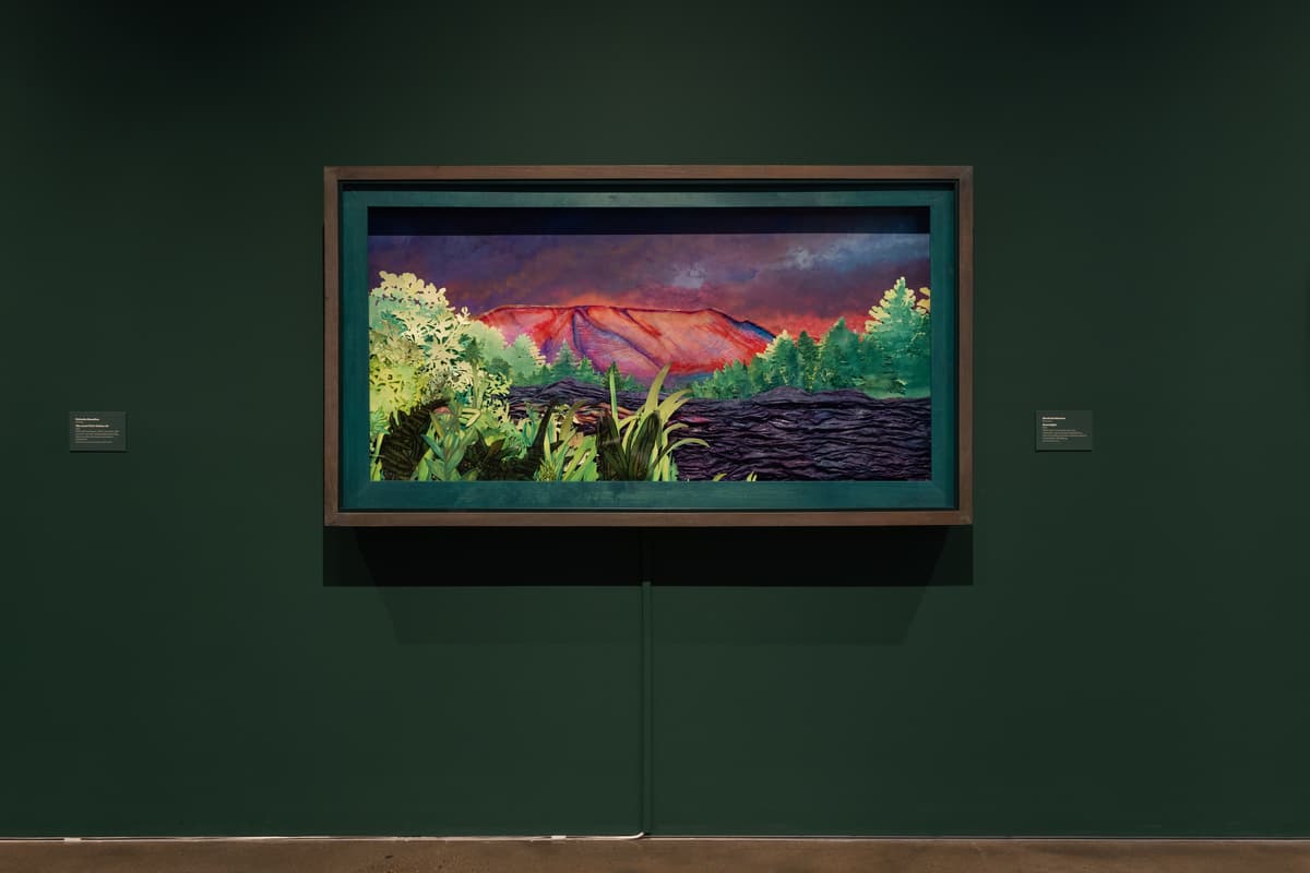 A wide, framed landscape depicting a vibrant red-orange mountain under a dark, moody purple sky. The foreground is filled with textured, layered cutouts of green trees and tall grass, giving the piece a three-dimensional, diorama-like quality. The piece is set within a deep, teal-painted inner frame and a larger wooden outer frame and hung on a dark green wall.