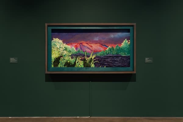 A wide, framed landscape depicting a vibrant red-orange mountain under a dark, moody purple sky. The foreground is filled with textured, layered cutouts of green trees and tall grass, giving the piece a three-dimensional, diorama-like quality. The piece is set within a deep, teal-painted inner frame and a larger wooden outer frame and hung on a dark green wall.