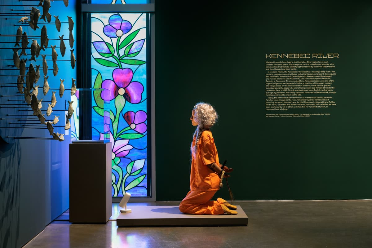 A sculpture of a figure with long grey hair, dressed in an orange jumpsuit, is kneeling on a low platform with their hands bound behind their back. To the right, a dark green wall displays a large text panel titled "KENNEBEC RIVER," while to the left, a tall stained-glass window with vibrant purple flowers and a blue background illuminates the scene. A portion of a circular wall sculpture featuring numerous small fish models is visible on the far left.