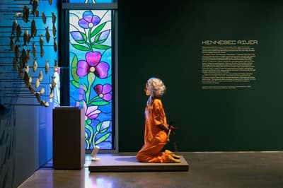 A sculpture of a figure with long grey hair, dressed in an orange jumpsuit, is kneeling on a low platform with their hands bound behind their back. To the right, a dark green wall displays a large text panel titled "KENNEBEC RIVER," while to the left, a tall stained-glass window with vibrant purple flowers and a blue background illuminates the scene. A portion of a circular wall sculpture featuring numerous small fish models is visible on the far left.