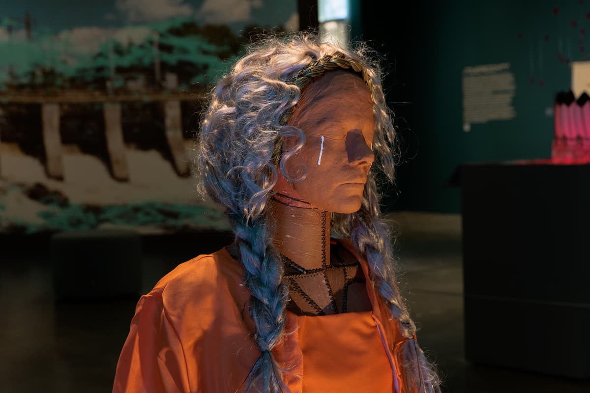 A close-up of a figurative sculpture in an orange jumpsuit with long, braided silver-blue hair and a clay-like face, with a single white tear painted on its cheek.