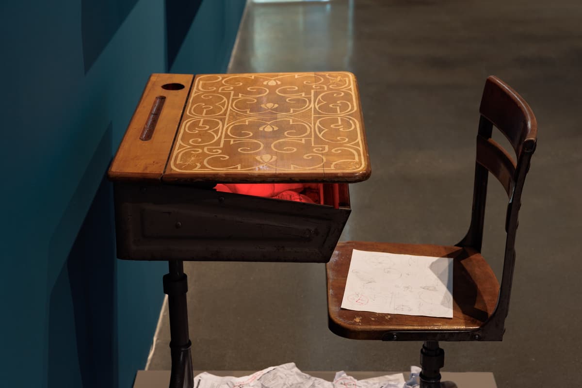 An installation comprising a vintage wooden school desk and chair. The desk is decorated with an intricate, light-colored scrollwork pattern and slightly propped open to reveal a glowing red light emanating from within the storage compartment. A piece of paper with hand-drawn sketches sits on the chair's seat, while crumpled papers are scattered on the floor beneath the desk.