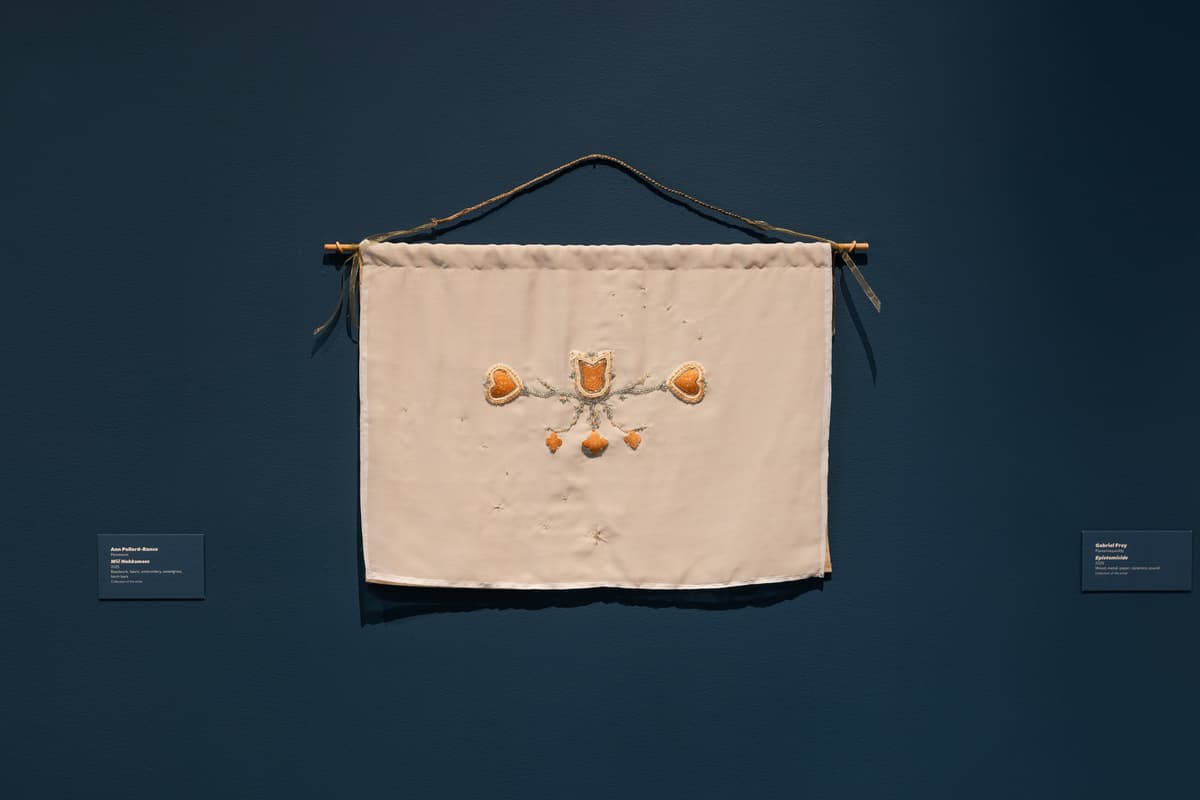 A cream-colored rectangular fabric banner hangs from a thin wooden dowel against a dark teal gallery wall. The center of the textile features delicate beadwork and embroidery, including three heart-like shapes and floral motifs in shades of amber and silver. The banner is suspended by a rustic, twisted cord.