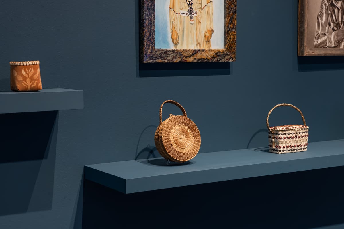 Three handcrafted baskets displayed on floating shelves. On the lower, longer shelf, a round, woven grass basket with a circular handle sits next to a rectangular basket featuring intricate white, red, and black woven patterns and a braided handle. To the left, a higher, smaller shelf holds a cylindrical vessel made of reddish-brown bark with a light-colored floral motif etched into its surface. Above the baskets, two framed works of art, each depicting a single figure, are partially visible.