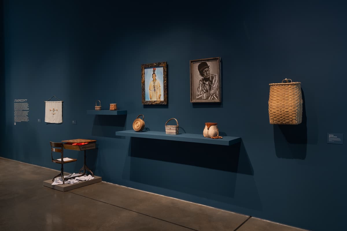 View of exhibition of contemporary Indigenous artwork in gallery with a dark teal wall and polished concrete floors. From left to right: a hanging textile work, a sculptural installation, several woven baskets on floating shelves, two framed works —one a vibrant painting of a figure in traditional regalia and the other a sepia-toned image of an elder. To the right, a large, tall utility basket is mounted directly to the wall.