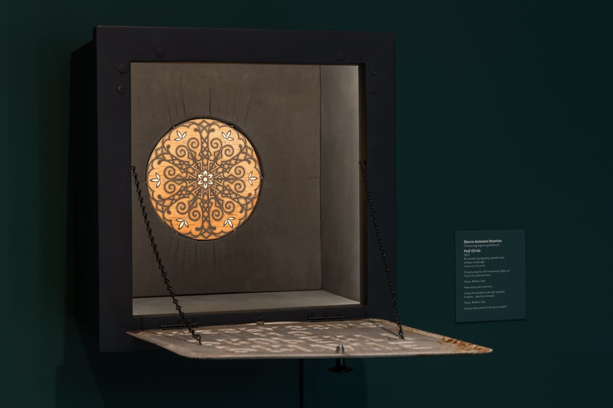 A black square shadow box is mounted on the wall with its front panel—a weathered, white metal "PRIVATE PROPERTY" sign—hinged open and held flat by thin black chains. Inside the illuminated box, a circular piece of light-toned birch bark is displayed, featuring an intricate, dark brown symmetrical mandala pattern created with pyrography and white highlights.