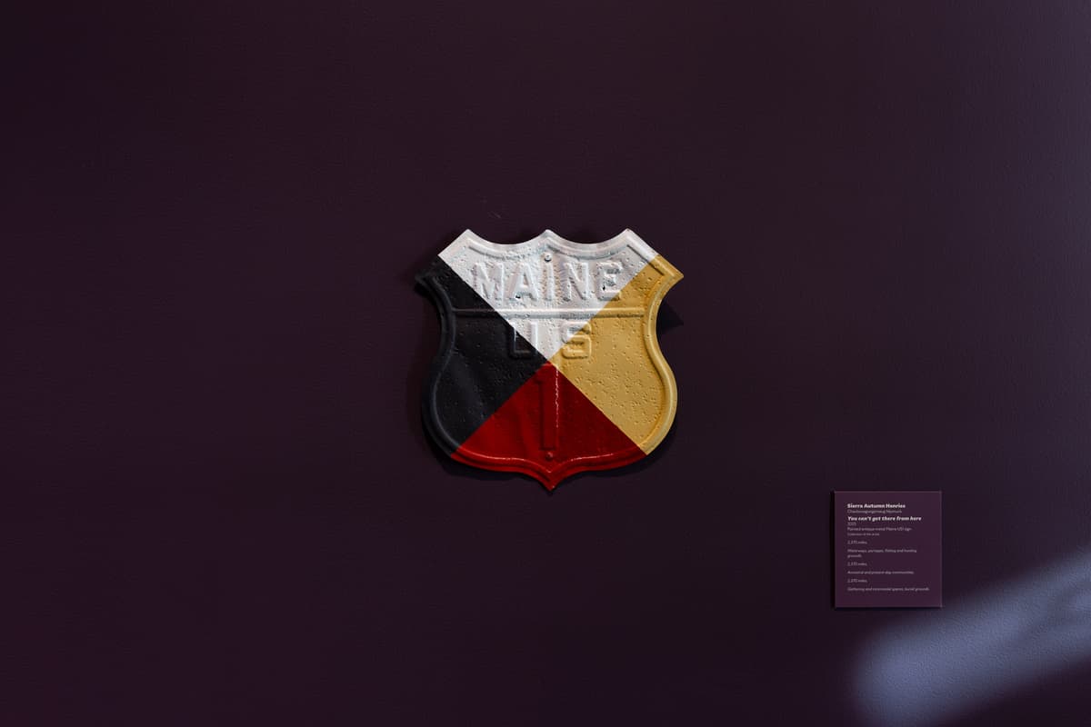 Against a dark plum wall, a weathered metal shield-shaped sign, an antique US Route 1 marker for Maine, divided into four distinct quadrants painted in the colors of a medicine wheel: white at the top, yellow on the right, red at the bottom, and black on the left.